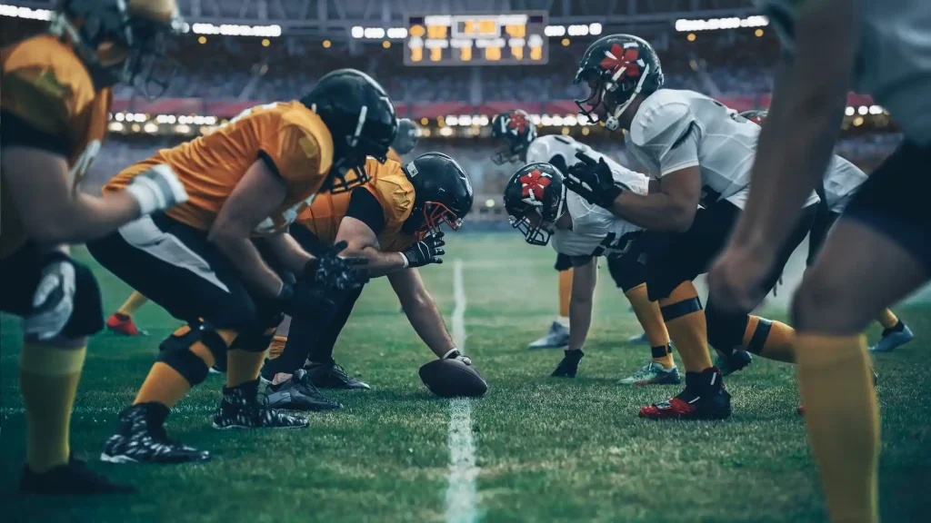 Zwei American Football Teams stehen sich auf Football Spielfeld gegenueber und bereiten sich auf den Angriff vor.