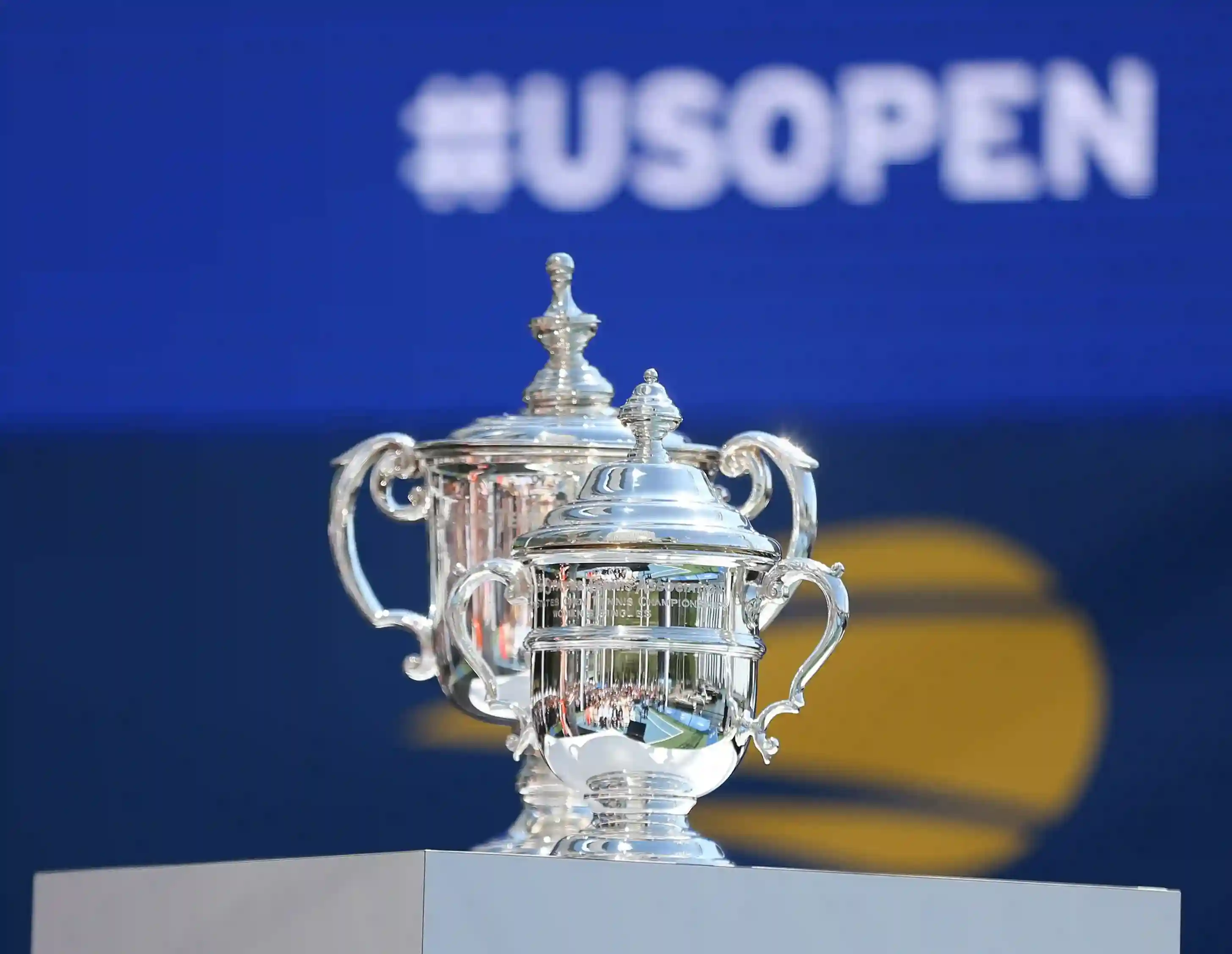 Die Trophäen der US Open vor einem blau-schwarzen Hintergrund mit der weißen Aufschrift „US OPEN”.
